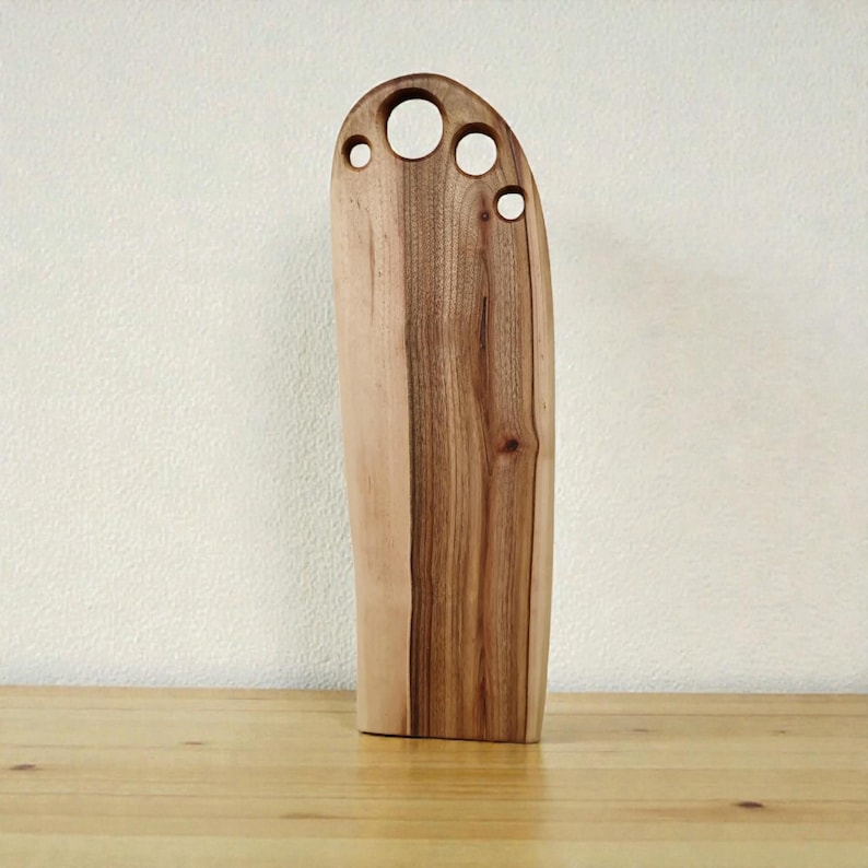 Pode incluir: Uma t&aacute;bua de corte de madeira alta e vertical com um padr&atilde;o de gr&atilde;o natural em tons de castanho claro e escuro. A extremidade superior &eacute; arredondada e apresenta quatro aberturas circulares de tamanhos variados numa fila horizontal.