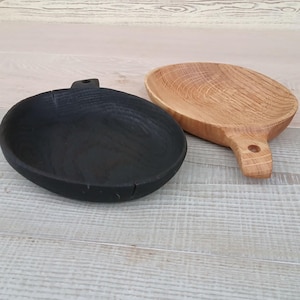 K&ouml;nnte beinhalten: Zwei Servierschalen aus Holz. Eine ist schwarz, die andere aus Naturholz. Beide haben einen Griff mit einem Loch. Die schwarze Schale hat eine glatte Oberfl&auml;che, w&auml;hrend die Naturschale die Holzmaserung zeigt.