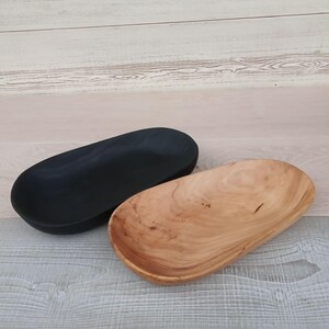 May include: Two wooden oval bowls. One is painted black, the other is natural wood with visible grain. The bowls are displayed on a light-colored wooden surface. These decorative bowls are suitable for serving or display.