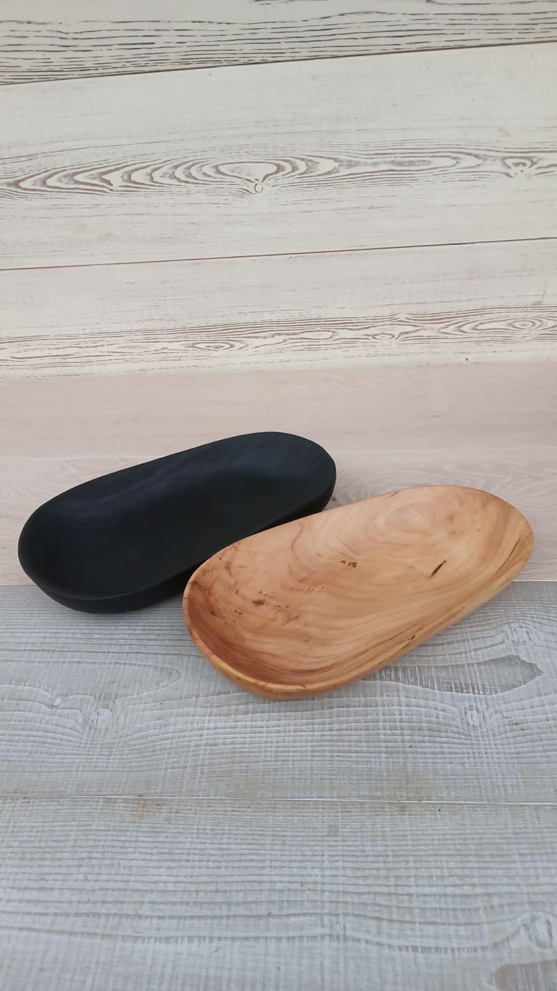 May include: Two wooden oval bowls. One is painted black, the other is natural wood with visible grain. The bowls are displayed on a light-colored wooden surface. These decorative bowls are suitable for serving or display.