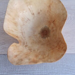 Op de afbeelding: Een handgemaakte houten kom met een unieke, organische vorm. De kom is gemaakt van lichtgekleurd hout, met natuurlijke nerfpatronen en een warme, aardse tint. De onregelmatige rand van de kom draagt bij aan de rustieke charme.