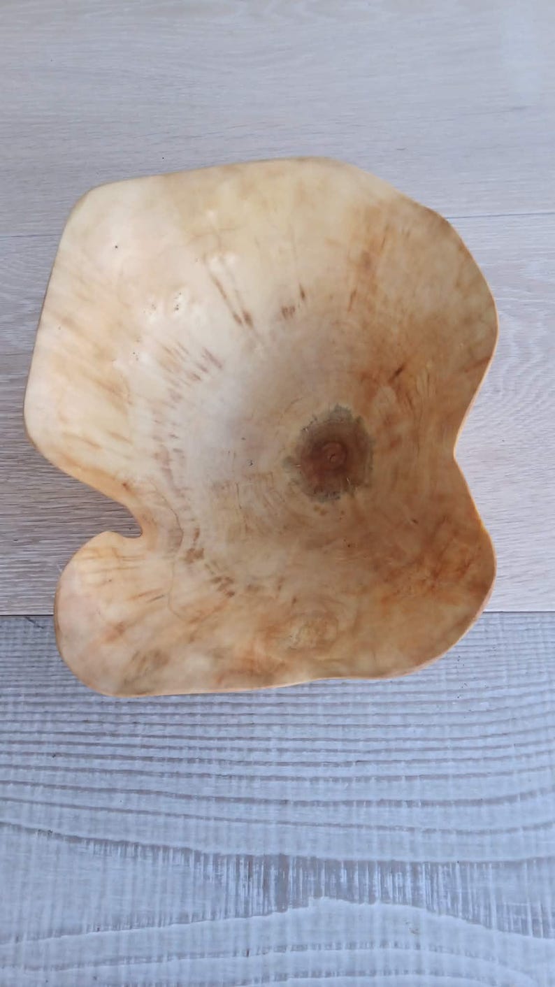Op de afbeelding: Een handgemaakte houten kom met een unieke, organische vorm. De kom is gemaakt van lichtgekleurd hout, met natuurlijke nerfpatronen en een warme, aardse tint. De onregelmatige rand van de kom draagt bij aan de rustieke charme.