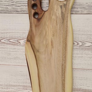 May include: A light brown wooden serving board with a natural live edge, featuring a large circular handle at the top and four smaller circular cutouts along the side. The wood grain displays varied tones of tan, cream, and dark brown throughout.