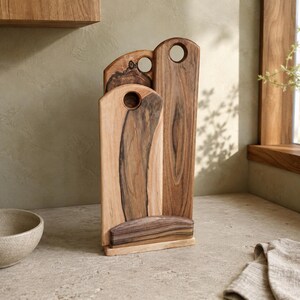 Op de afbeelding: Een set van drie verticale houten snijplanken met natuurlijke houtnerven, uitgevoerd in donkerbruine en lichtbruine tinten. Elke plank heeft een ronde uitsparing aan de bovenkant om op te hangen en ze staan in een houten standaard.
