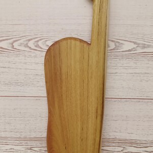 May include: A light brown wooden cutting board with a smooth, elongated rectangular body and a curved side edge. The board features a long, narrow handle extending from the top, which includes a circular hole for hanging or storage purposes.