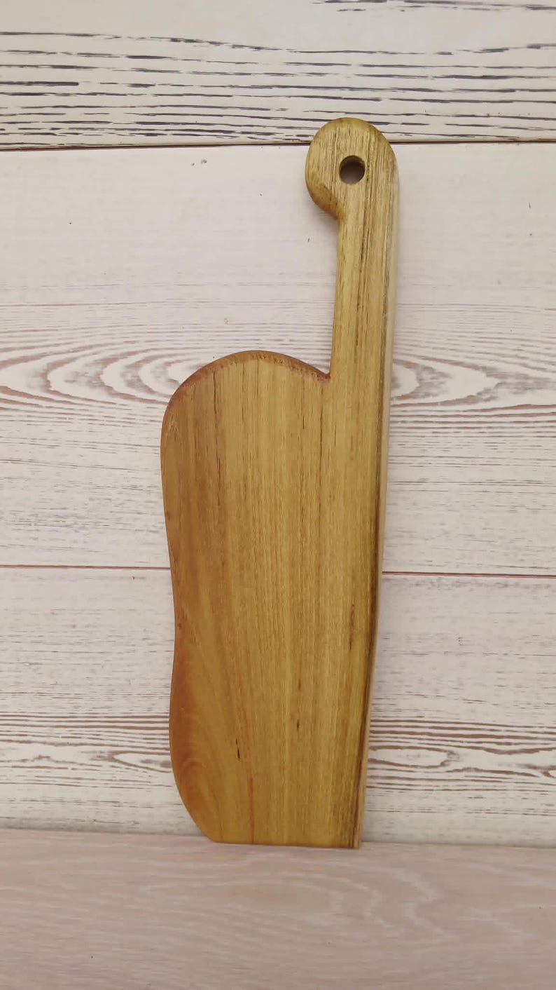 May include: A light brown wooden cutting board with a smooth, elongated rectangular body and a curved side edge. The board features a long, narrow handle extending from the top, which includes a circular hole for hanging or storage purposes.
