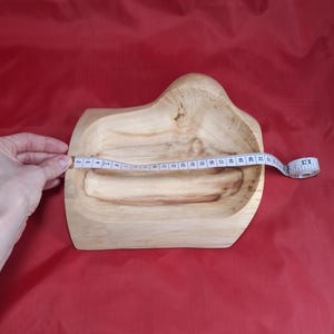 May include: A light-coloured wooden bowl with an unusual, organic shape. The bowl is being measured with a tape measure, showing a length of approximately 33 cm. The bowl sits on a red surface.