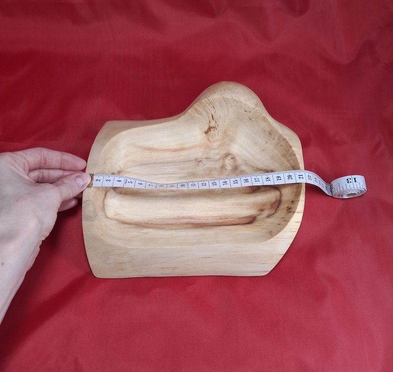 May include: A light-coloured wooden bowl with an unusual, organic shape. The bowl is being measured with a tape measure, showing a length of approximately 33 cm. The bowl sits on a red surface.