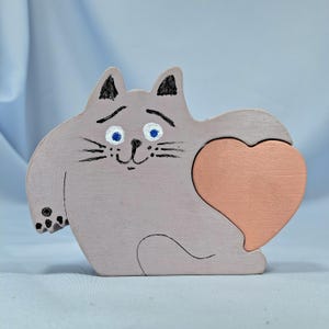 以下が含まれることがあります： ハート型のピースが付いた、猫の形をした木製パズル。猫は薄いグレーで塗装されており、黒いひげ、耳、足のディテールがあります。ハートはソフトピーチ色で、猫は青い目をしています。