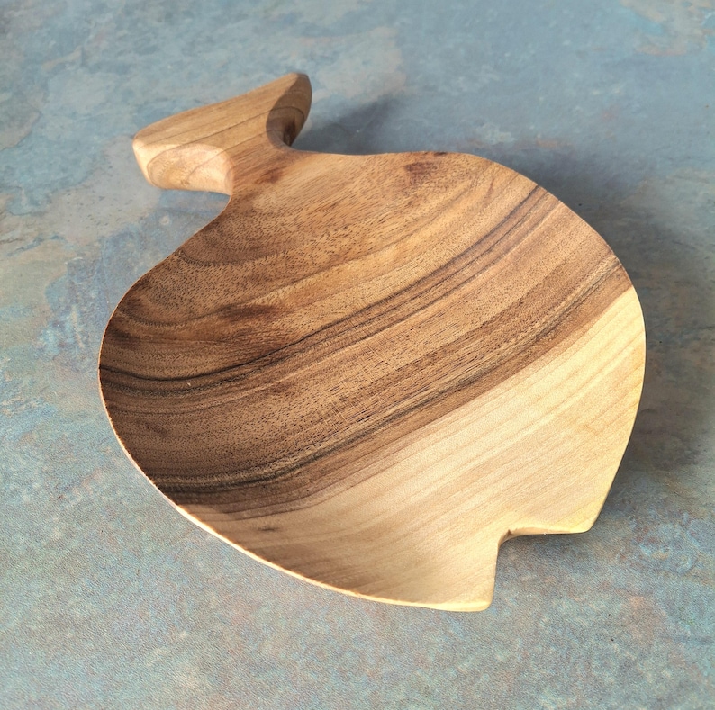May include: A wooden serving bowl with an unusual, organic form. The bowl has a natural wood grain and a handle-like extension. The bowl is approximately 30 cm long and 20 cm wide.