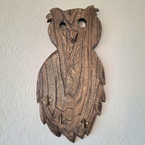 May include: Wooden owl-shaped key holder with a natural wood grain finish. The key holder has three brass-colored hooks at the bottom and two eye holes. The owl's head has a textured appearance, and the overall design is rustic.