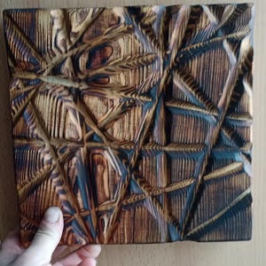 May include: A square, wooden wall art piece with a geometric, carved design. The artwork features a complex pattern of intersecting lines and shapes, with a mix of light and dark brown tones. The piece is held by a hand.