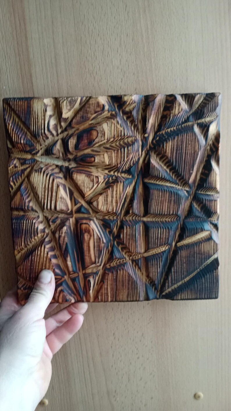 May include: A square, wooden wall art piece with a geometric, carved design. The artwork features a complex pattern of intersecting lines and shapes, with a mix of light and dark brown tones. The piece is held by a hand.