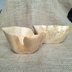 Op de afbeelding: Twee houten kommen met een natuurlijke, lichtgeleurde afwerking. De kommen hebben organische, asymmetrische vormen en staan tegen een neutrale achtergrond. De houtnerf is zichtbaar, wat bijdraagt aan de rustieke esthetiek. Deze kommen zijn ideaal om te serveren of tentoon te stellen.