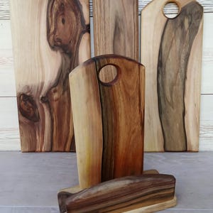Op de afbeelding: Een verzameling van vier houten snijplanken met natuurlijke houtnerven en boomkanten. Drie planken staan rechtop, elk met een rond handvatgat aan de bovenkant, terwijl &eacute;&eacute;n plank horizontaal in een houten standaard rust.
