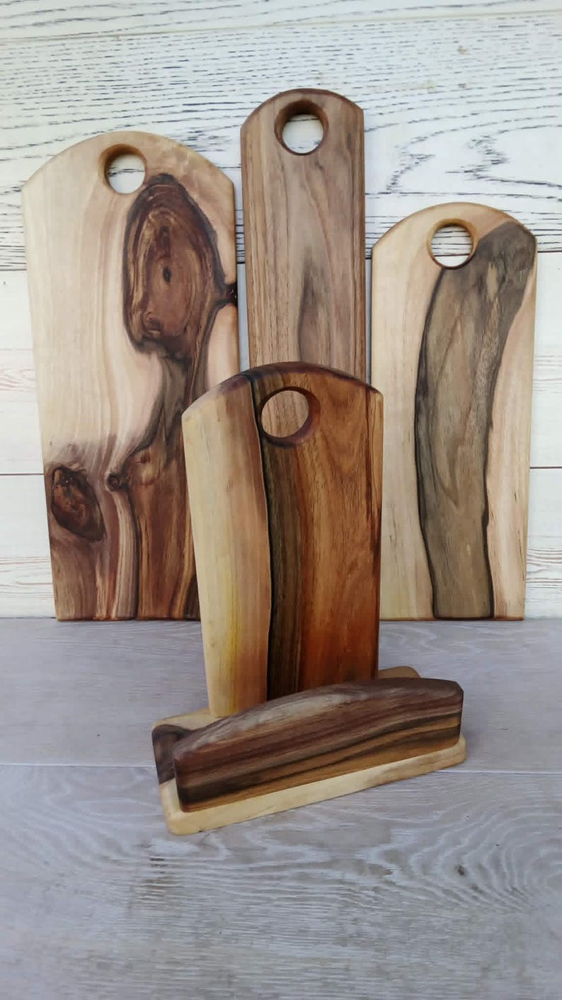 Op de afbeelding: Een verzameling van vier houten snijplanken met natuurlijke houtnerven en boomkanten. Drie planken staan rechtop, elk met een rond handvatgat aan de bovenkant, terwijl &eacute;&eacute;n plank horizontaal in een houten standaard rust.