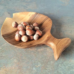 May include: A wooden serving dish shaped like a fish, holding a collection of hazelnuts. The dish is crafted from light brown wood, showcasing a natural grain. The hazelnuts are brown with a pale shell. The background is a mottled blue and grey.