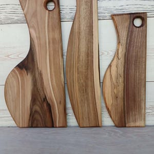 Op de afbeelding: Drie houten serveerplanken met natuurlijke houtnerven en boomkanten. Elke plank heeft een uitgesproken, langwerpige vorm met een rond gat aan de bovenkant om op te hangen. Het hout vertoont gevarieerde bruintinten van licht tot donker.