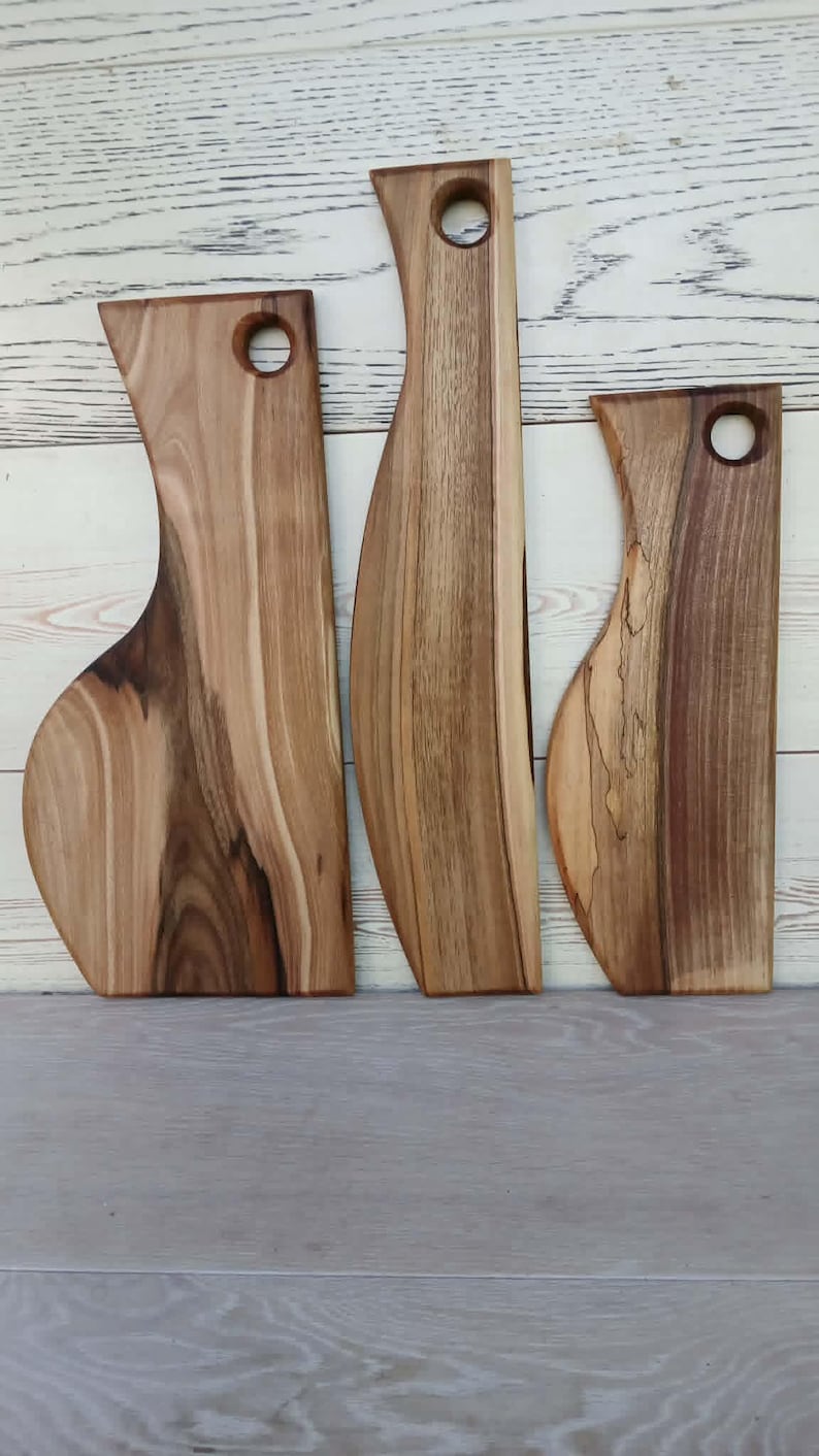 Op de afbeelding: Drie houten serveerplanken met natuurlijke houtnerven en boomkanten. Elke plank heeft een uitgesproken, langwerpige vorm met een rond gat aan de bovenkant om op te hangen. Het hout vertoont gevarieerde bruintinten van licht tot donker.