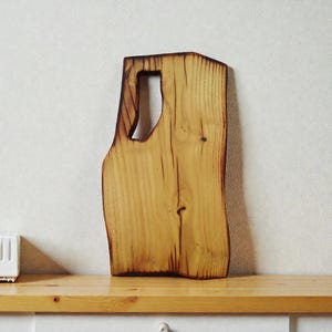Pode incluir: Uma t&aacute;bua de corte de madeira retangular com uma borda natural e um acabamento escuro carbonizado nas laterais. A t&aacute;bua apresenta um padr&atilde;o de veios de madeira vertical proeminente, um pequeno n&oacute; e uma al&ccedil;a retangular recortada no topo.
