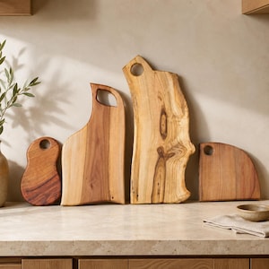 Puede incluir: Cuatro tablas de cortar de madera de diversas formas y tama&ntilde;os, cada una con un borde natural y un orificio circular para colgar. Las tablas muestran patrones de veta de madera distintos en tonos beige claro, marr&oacute;n dorado y marr&oacute;n rojizo.