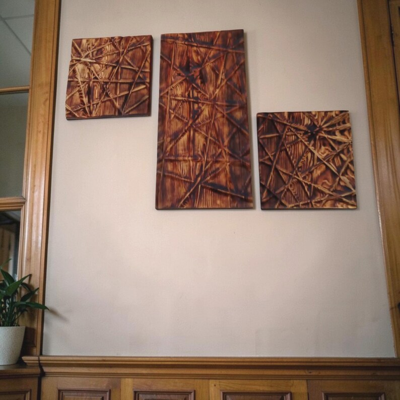 May include: Three wooden wall art pieces with a geometric design. The art features a network of intersecting lines in a warm brown hue, creating a textured, abstract pattern. The pieces are rectangular and square, mounted on a neutral-coloured wall.