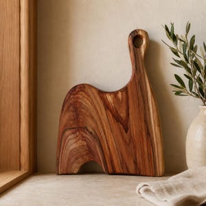 May include: A wooden cutting board with a natural grain pattern, featuring a curved silhouette and an integrated circular handle hole at the top. The board displays rich brown tones with darker streaks throughout its smooth, polished surface.