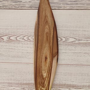 May include: A long, narrow wooden serving board with a tapered, paddle-like shape and a rounded handle featuring a circular hole at the top. The board displays a prominent, swirling natural wood grain pattern in shades of light and dark brown.