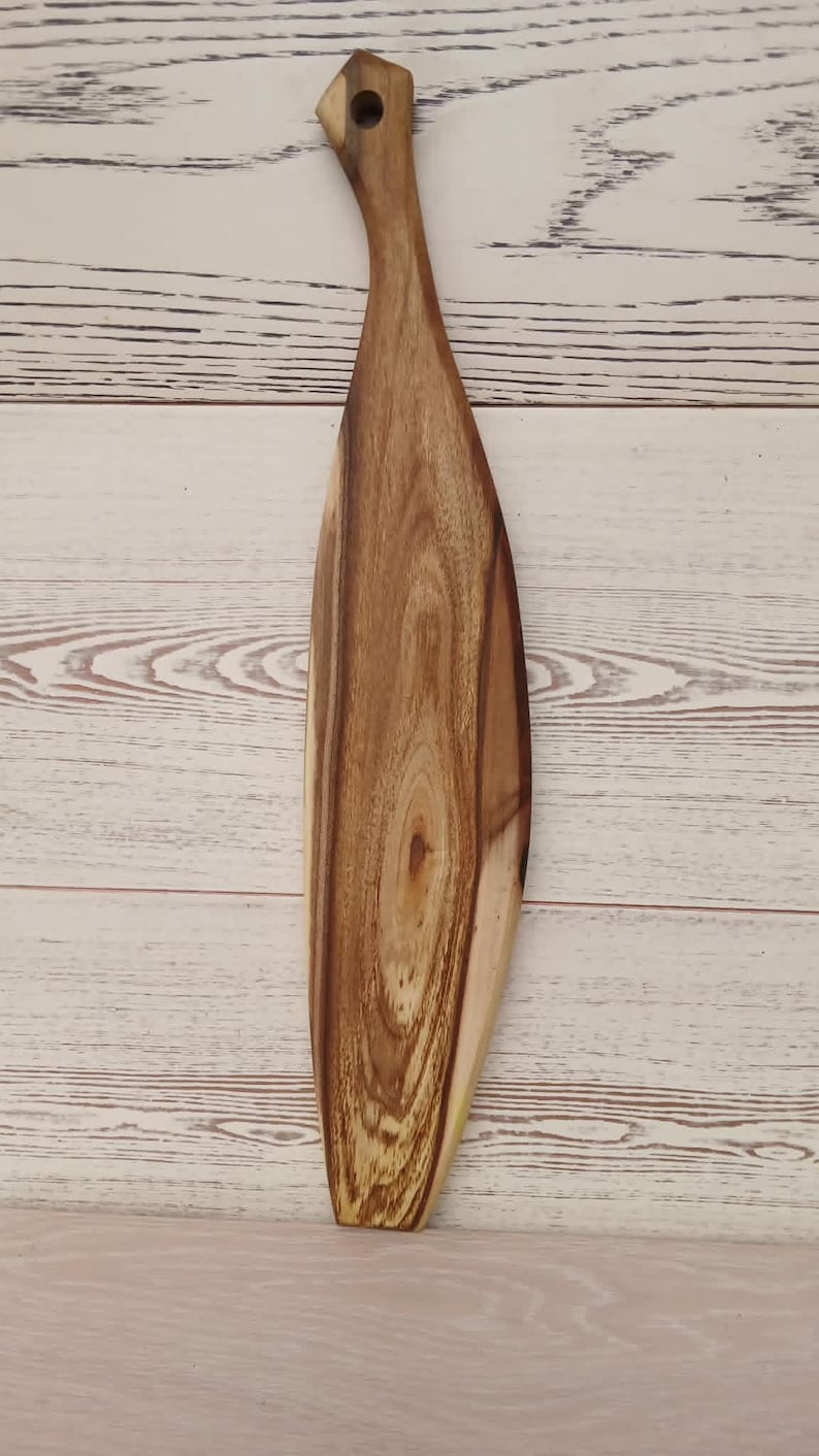 May include: A long, narrow wooden serving board with a tapered, paddle-like shape and a rounded handle featuring a circular hole at the top. The board displays a prominent, swirling natural wood grain pattern in shades of light and dark brown.