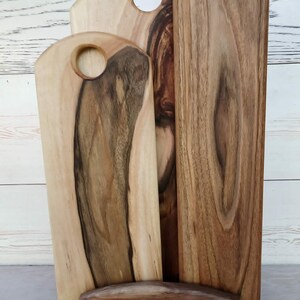 Op de afbeelding: Drie rechthoekige houten serveerplanken met afgeronde bovenkanten en ronde handvatopeningen, geplaatst in een houten standaard. De planken vertonen natuurlijke houtnerven met contrasterende lichte en donkerbruine strepen door het hout.