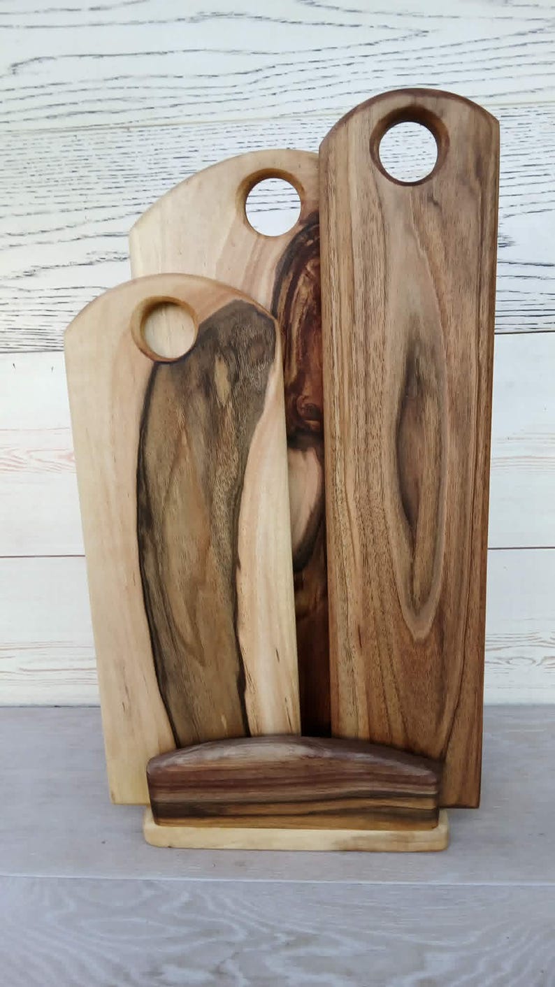 Op de afbeelding: Drie rechthoekige houten serveerplanken met afgeronde bovenkanten en ronde handvatopeningen, geplaatst in een houten standaard. De planken vertonen natuurlijke houtnerven met contrasterende lichte en donkerbruine strepen door het hout.