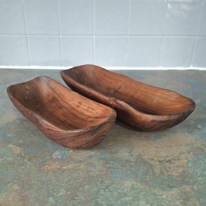 May include: Two wooden serving bowls with a natural, dark brown finish. The bowls have a long, boat-like shape with rounded edges and visible wood grain. They are placed on a mottled green and blue surface.