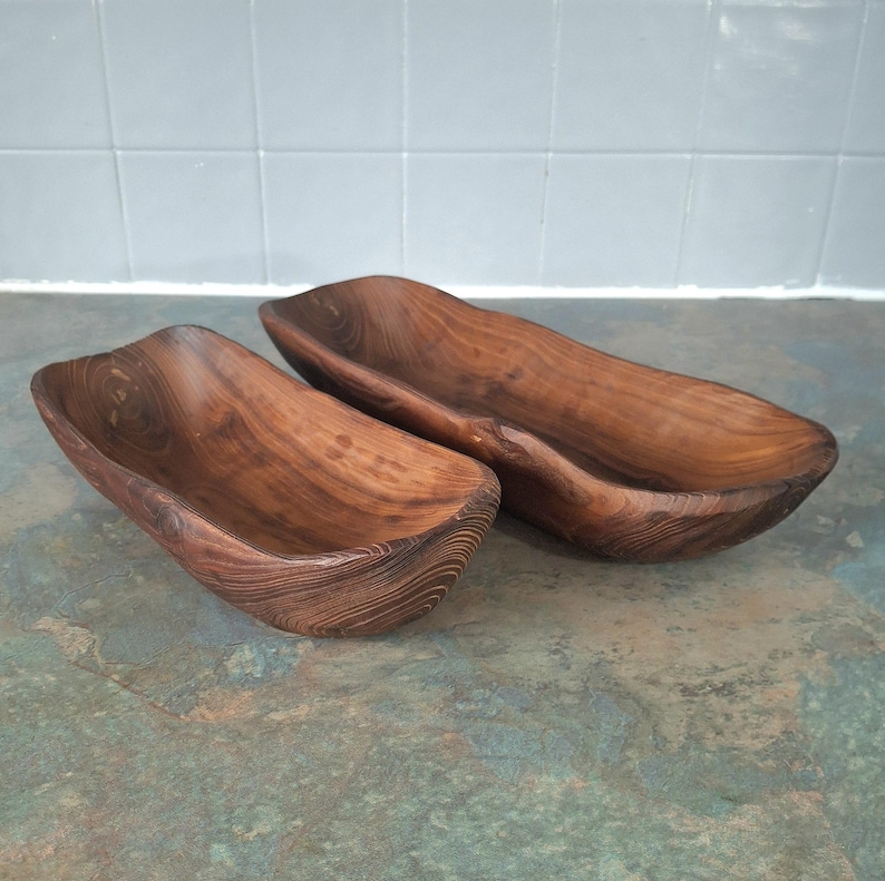 May include: Two wooden serving bowls with a natural, dark brown finish. The bowls have a long, boat-like shape with rounded edges and visible wood grain. They are placed on a mottled green and blue surface.