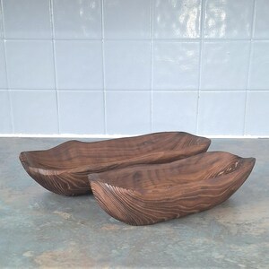 May include: Two wooden serving bowls with a natural wood grain finish. The bowls have a long, curved shape and are made of dark brown wood. The bowls are likely handmade and suitable for serving food or as decorative pieces.