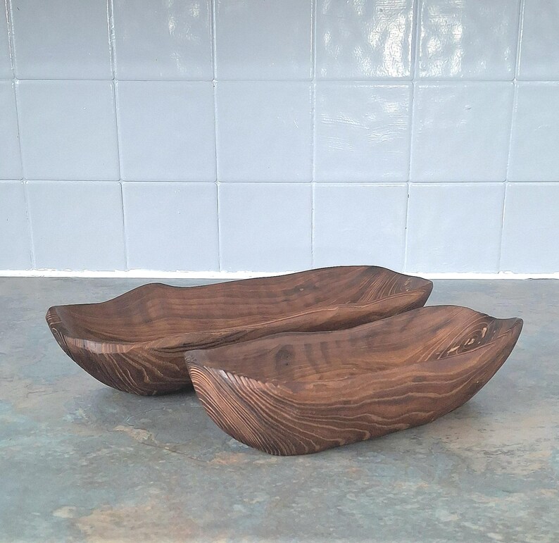 May include: Two wooden serving bowls with a natural wood grain finish. The bowls have a long, curved shape and are made of dark brown wood. The bowls are likely handmade and suitable for serving food or as decorative pieces.