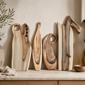 May include: Four wooden chopping boards with natural grain patterns and dark brown streaks. The boards feature varied shapes, including one with a circular handle, one with four small holes, one narrow tapered board, and one with an angled top handle.
