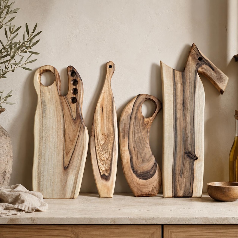 May include: Four wooden chopping boards with natural grain patterns and dark brown streaks. The boards feature varied shapes, including one with a circular handle, one with four small holes, one narrow tapered board, and one with an angled top handle.