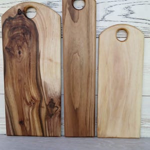 Op de afbeelding: Drie rechthoekige houten snijplanken van verschillende breedtes, elk met een afgeronde bovenkant en een cirkelvormig handvat. De planken vertonen natuurlijke houtnerven in tinten van lichtbruin, middenbruin en donker chocoladebruin.