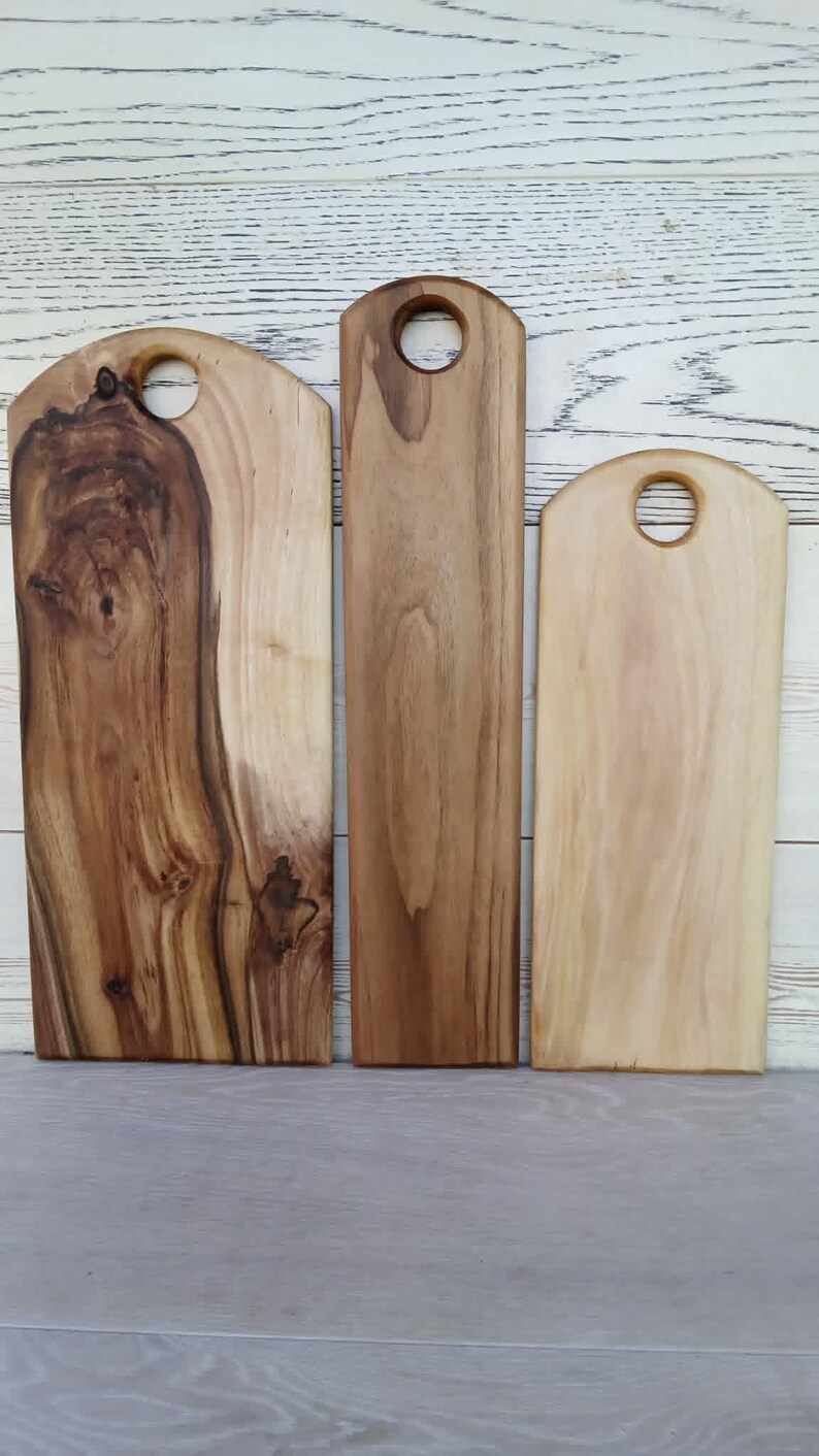 Op de afbeelding: Drie rechthoekige houten snijplanken van verschillende breedtes, elk met een afgeronde bovenkant en een cirkelvormig handvat. De planken vertonen natuurlijke houtnerven in tinten van lichtbruin, middenbruin en donker chocoladebruin.