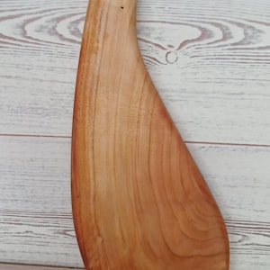 May include: A light brown wooden cutting board with a smooth, curved, asymmetrical shape and a circular hole near the top handle section. The wood grain is visible throughout the surface, showing natural variations in color and texture.
