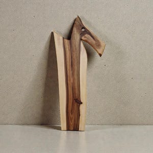 May include: A wooden serving board featuring a natural live edge and a distinct, elongated shape that resembles a stylised animal head. The board displays a prominent grain pattern with contrasting light cream and deep brown wood tones.
