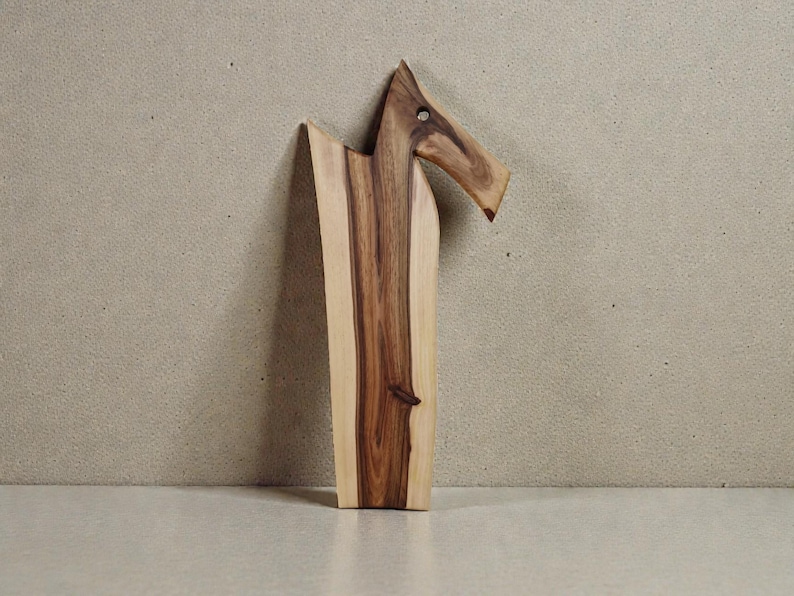 May include: A wooden serving board featuring a natural live edge and a distinct, elongated shape that resembles a stylised animal head. The board displays a prominent grain pattern with contrasting light cream and deep brown wood tones.