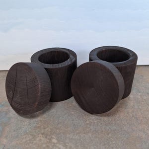 May include: Four dark brown wooden containers with lids. Two are cylindrical with a circular opening, and two are round with a flat top. The wood grain is visible, and the containers are arranged on a neutral surface.