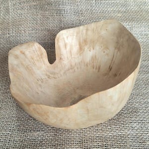 Op de afbeelding: Een handgemaakte houten kom met een lichte kleur en een organische vorm. De kom heeft een opvallende uitsparing en toont natuurlijke houtnerfpatronen. Geschikt voor serveren of decoratie. De diameter is ongeveer 25 cm.