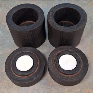 May include: Four dark stained wooden candle holders. Two are cylindrical with a hollow centre, and two are round with a recessed area holding a white tea light candle. The wood grain is visible.