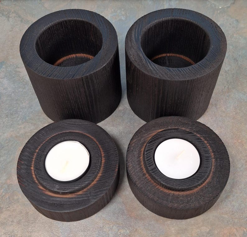 May include: Four dark stained wooden candle holders. Two are cylindrical with a hollow centre, and two are round with a recessed area holding a white tea light candle. The wood grain is visible.
