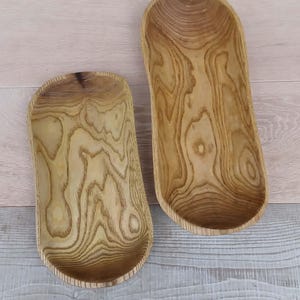 Op de afbeelding: Twee ovaalvormige houten kommen met een natuurlijke lichtbruine afwerking en zichtbare donkere houtnerfpatronen. De kommen zijn langwerpig met gladde, gebogen randen en een diep, hol midden, wat de organische textuur van het houtmateriaal laat zien.