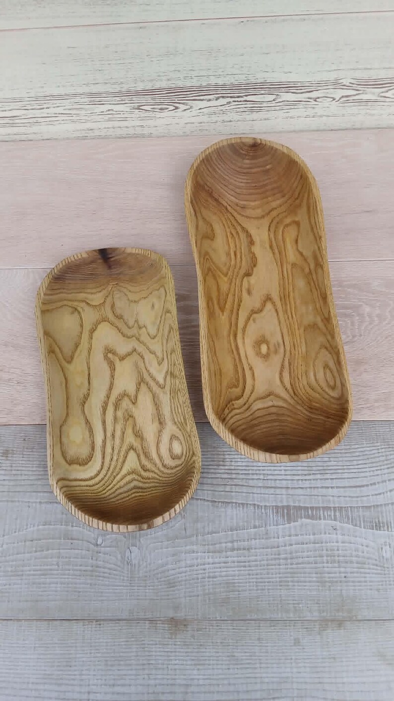 Op de afbeelding: Twee ovaalvormige houten kommen met een natuurlijke lichtbruine afwerking en zichtbare donkere houtnerfpatronen. De kommen zijn langwerpig met gladde, gebogen randen en een diep, hol midden, wat de organische textuur van het houtmateriaal laat zien.