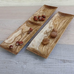 May include: Two elongated, rectangular wooden trays with a prominent light and dark brown wood grain pattern. The left tray contains several small brown hazelnuts, while the right tray holds three larger, textured brown walnuts.
