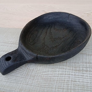 K&ouml;nnte beinhalten: Ein handgefertigter, schwarzer Holzservierl&ouml;ffel mit Griff und rundem Loch. Der L&ouml;ffel hat eine tiefe Sch&uuml;ssel und ist aus dunklem Holz gefertigt. Der L&ouml;ffel liegt auf einer hellen Holzoberfl&auml;che.